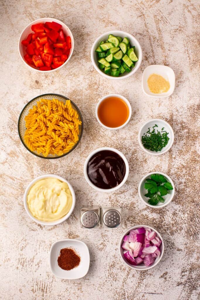 Ingredients for creamy BBQ pasta salad arranged in small bowls including rotini pasta, mayonnaise, BBQ sauce, apple cider vinegar, red bell pepper, English cucumber, red onion, fresh chives, parsley, garlic powder, and chili powder