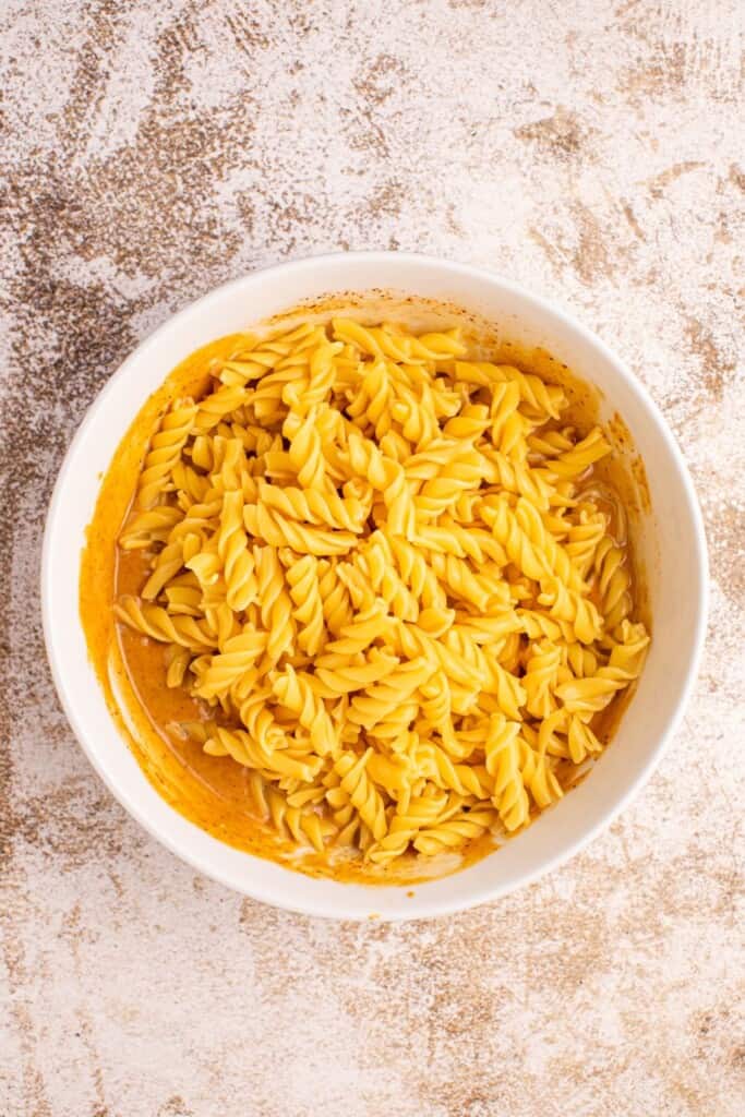 Cooked rotini pasta added to creamy BBQ dressing in a large mixing bowl, ready to be combined with fresh vegetables