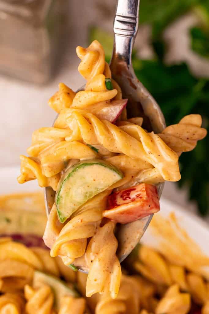 A spoonful of creamy BBQ pasta salad lifted from the serving bowl showing rotini, cucumber, red bell pepper, and red onion coated in smoky BBQ dressing