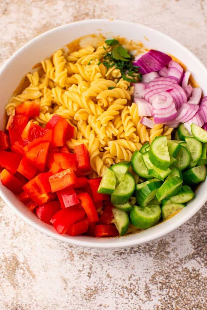 Rotini pasta, diced red bell pepper, sliced English cucumber, red onion, and fresh herbs arranged in a mixing bowl before tossing into creamy BBQ pasta salad