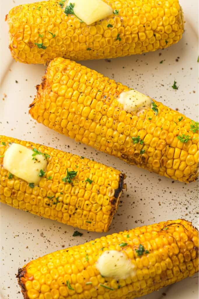 charred corn on the cob on a white plate. corn has melted pats of butter, flaky salt and sprinkles of parsley on tp