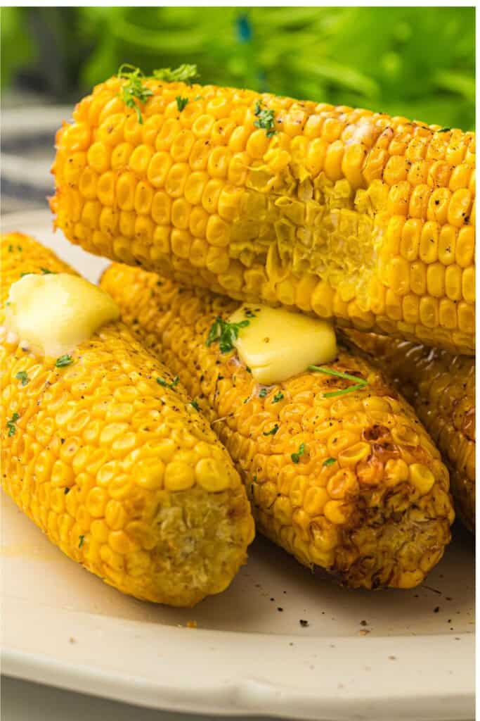 corn on cob stacked on top of each other, one ear has a bite out of it, the ther two ears have melted pats of butter wuth parsley sprinkled on top