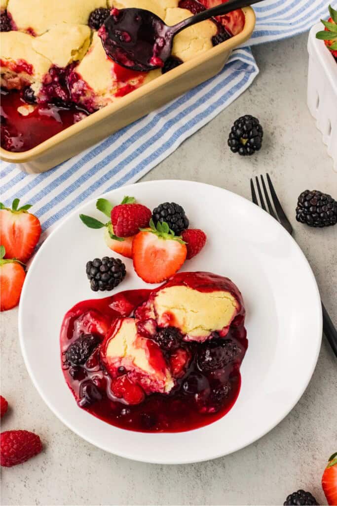 Slice of vegan berry cobbler on a white plate with fresh strawberries, blackberries, and raspberries