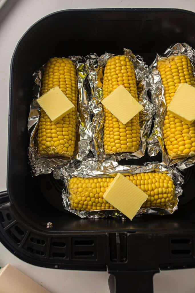 Overhead view of uncooked corn with pats of butter on top on foil in air fryer basket