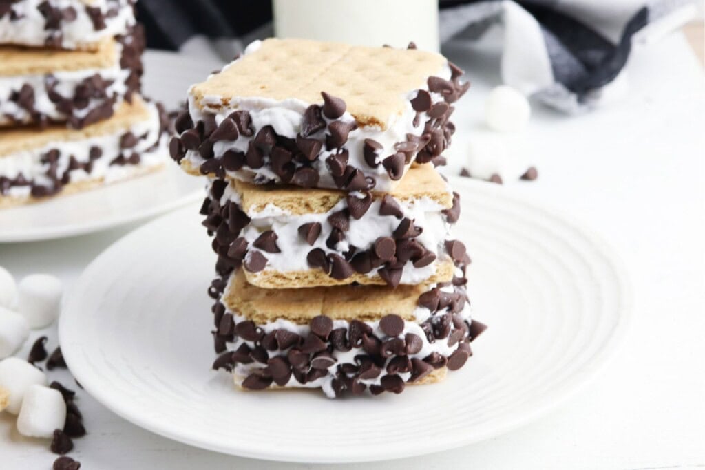 Frozen s'mores stacked on white plates with a bottle of milk, chocolate chips, and marshmallows.