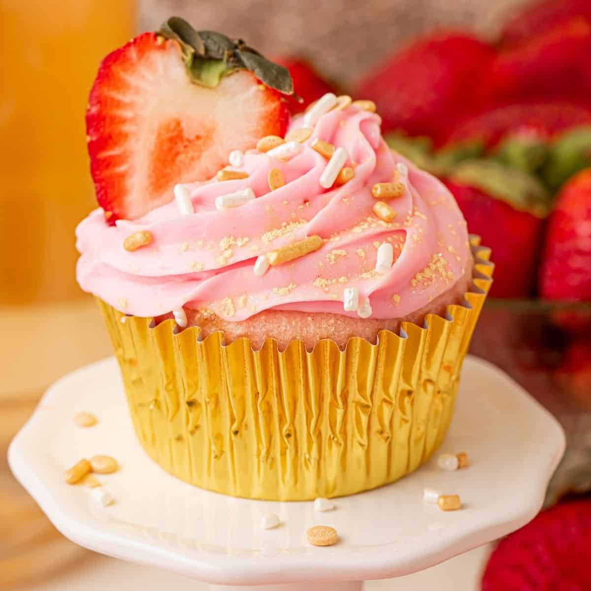 Pink champagne cupcake with strawberry slice in gold foil liner on cake stand