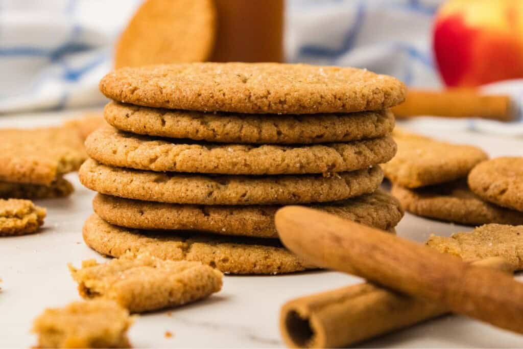 Apple cider snickerdoodles stacked on marble with a bottle of cider, a fresh apple and cinnamon sticks 