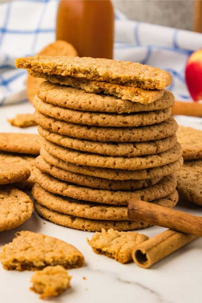 Tall stack of brown butter apple cider snickerdoodles with a broken cookie on top, cinnamon sticks and a bottle of cider behind