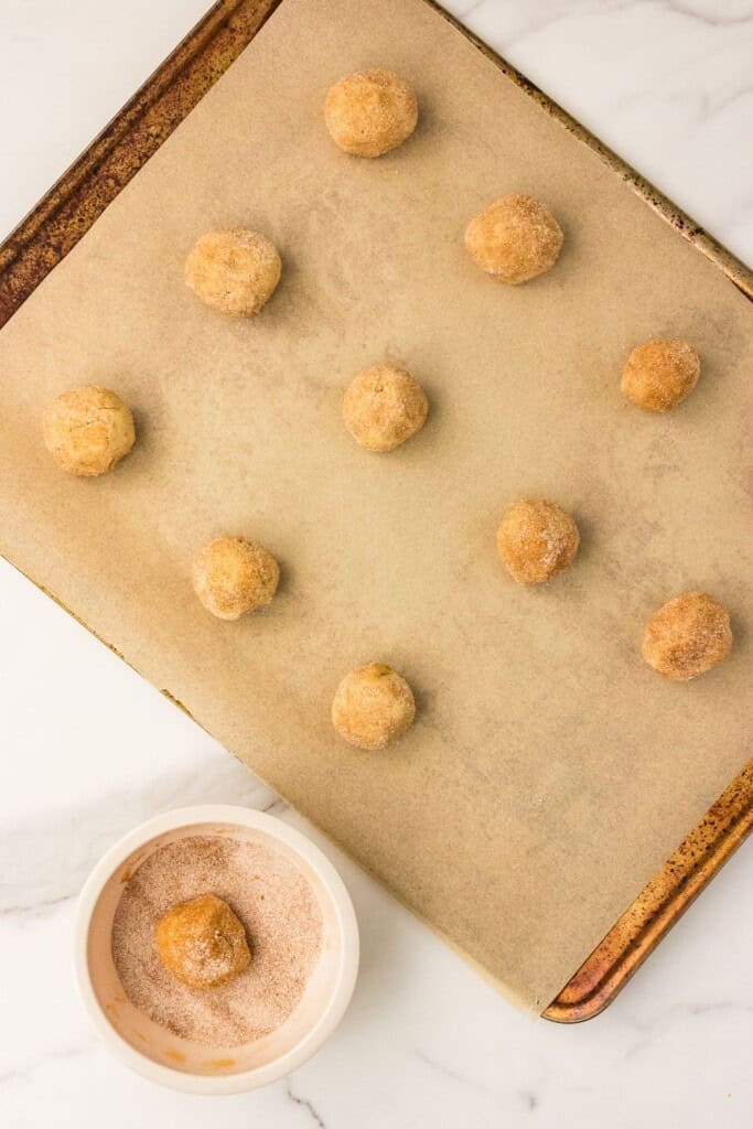 Snickerdoodle dough ball rolled in cinnamon sugar beside a parchment-lined baking sheet