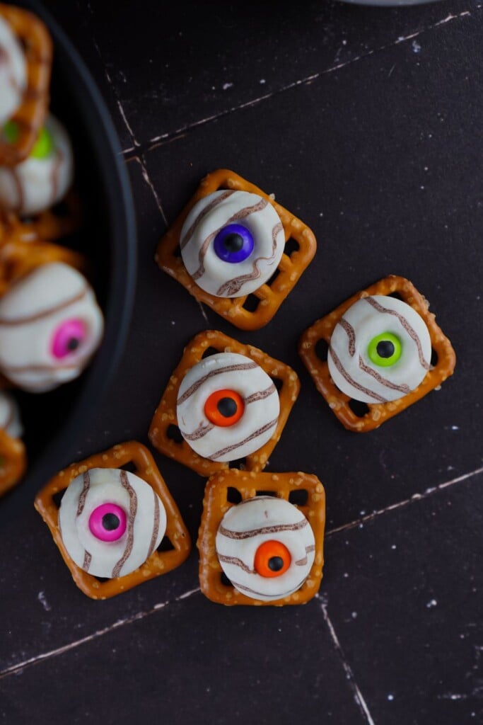 Five candy eye pretzel bites arranged on a dark surface next to a bowl of more bites