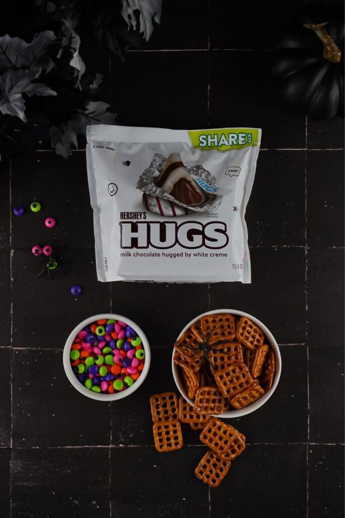 Bag of Hershey Hugs with bowls of pretzel snaps and colorful candy eyes on a dark table